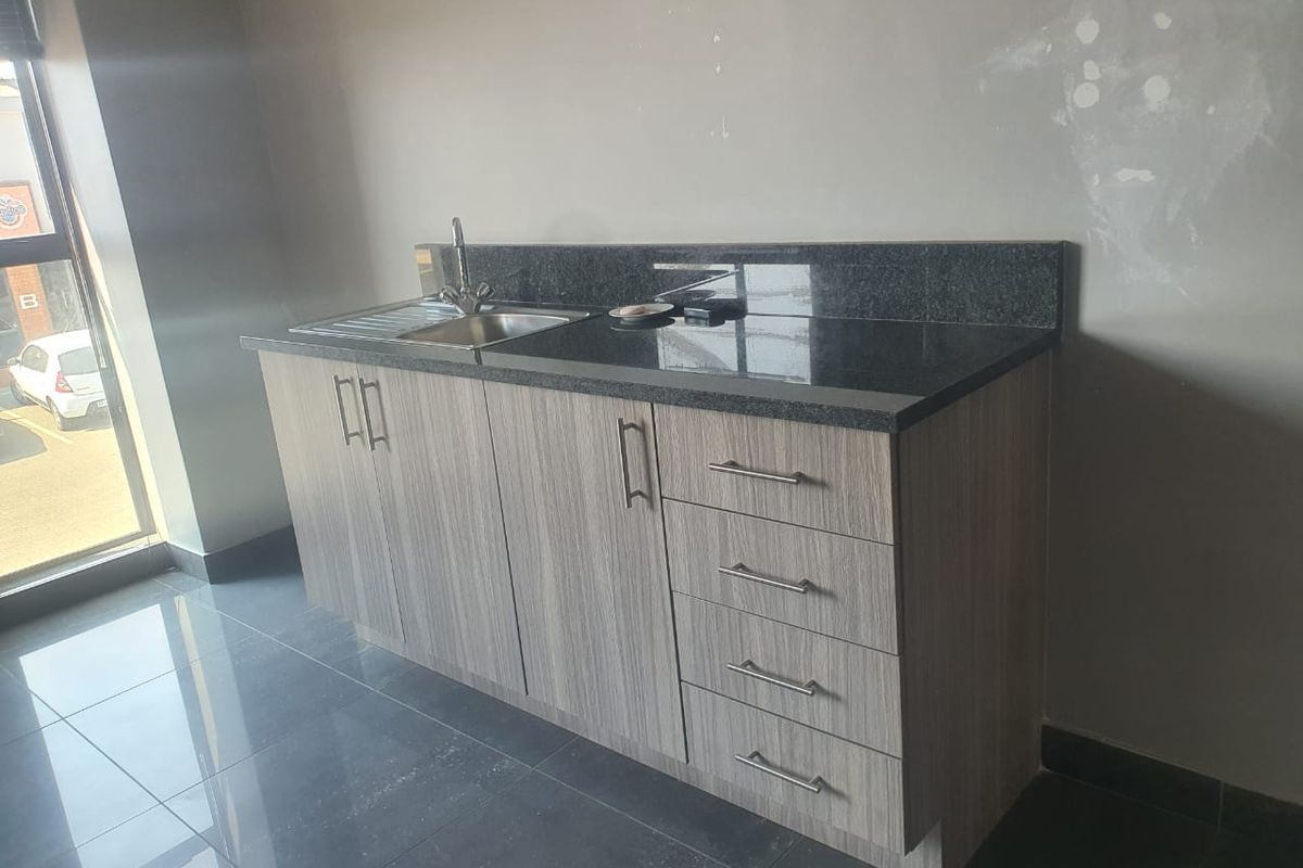 A compact kitchenette area featuring wood-grain cabinetry, a dark granite countertop, and a stainless steel sink, set against a tiled floor.