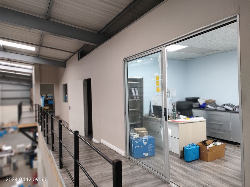 An office space located on a mezzanine level, featuring a sliding glass door, laminate flooring, and an exposed industrial steel roof structure.