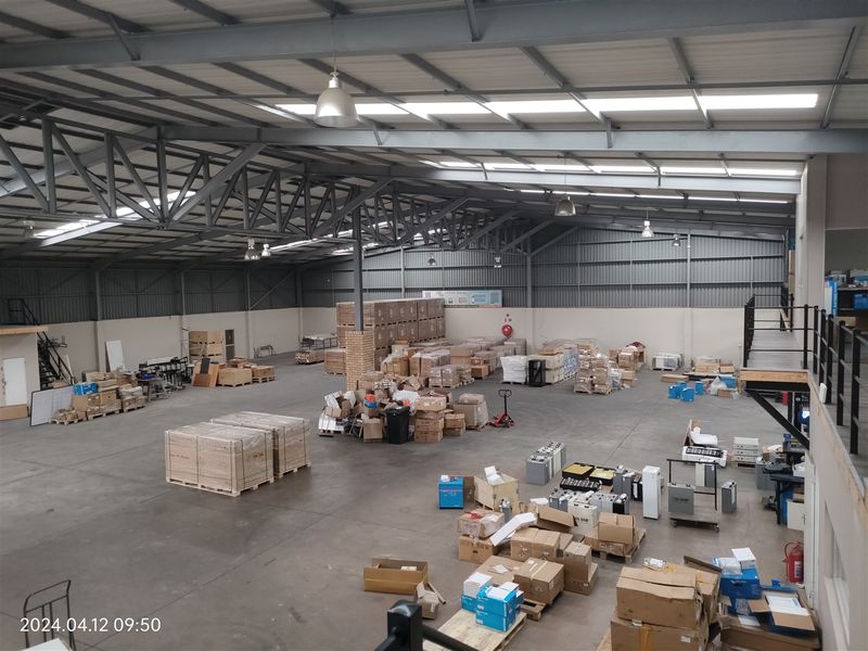 Spacious industrial warehouse featuring high ceilings with exposed steel trusses, concrete flooring, natural light via skylights, and an integrated mezzanine level.