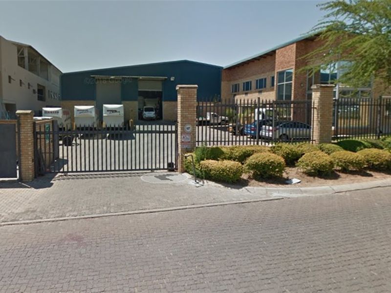 Front view of an industrial property featuring a secure gated entrance, paved driveway, and warehouse facility with roller shutter access.