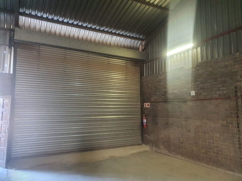 Industrial utility space featuring a large roller shutter door, exposed brick walls, and high corrugated metal roofing with fixed lighting.