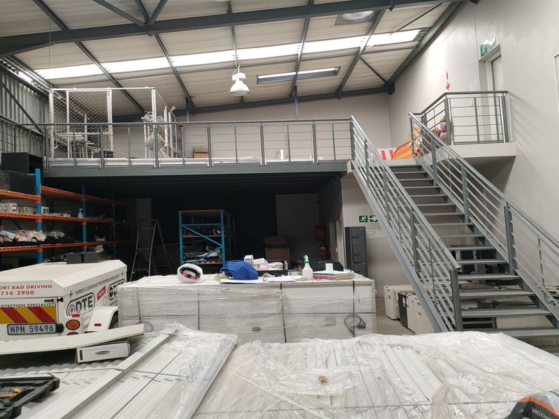 Industrial warehouse space featuring a mezzanine level with steel staircase, high ceilings with skylights, and functional lighting, suitable for storage or workshop operations.