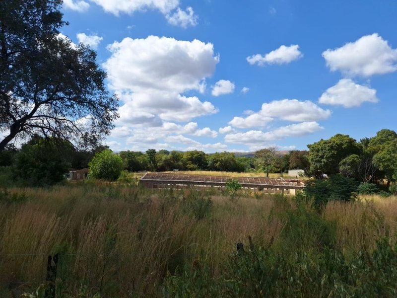 A wide view of a farm property featuring an outbuilding structure set within an expansive, overgrown field under a clear blue sky.