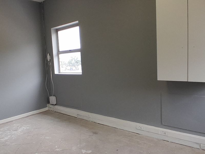 An empty office space featuring grey walls, a wall-mounted storage cabinet, and visible electrical trunking along the baseboards.
