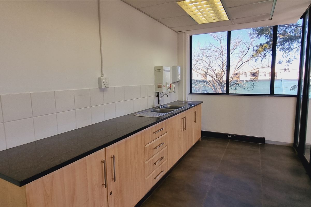 A functional office kitchenette featuring light wood cabinetry, a dark countertop, a stainless steel sink, and a wall-mounted water heater.