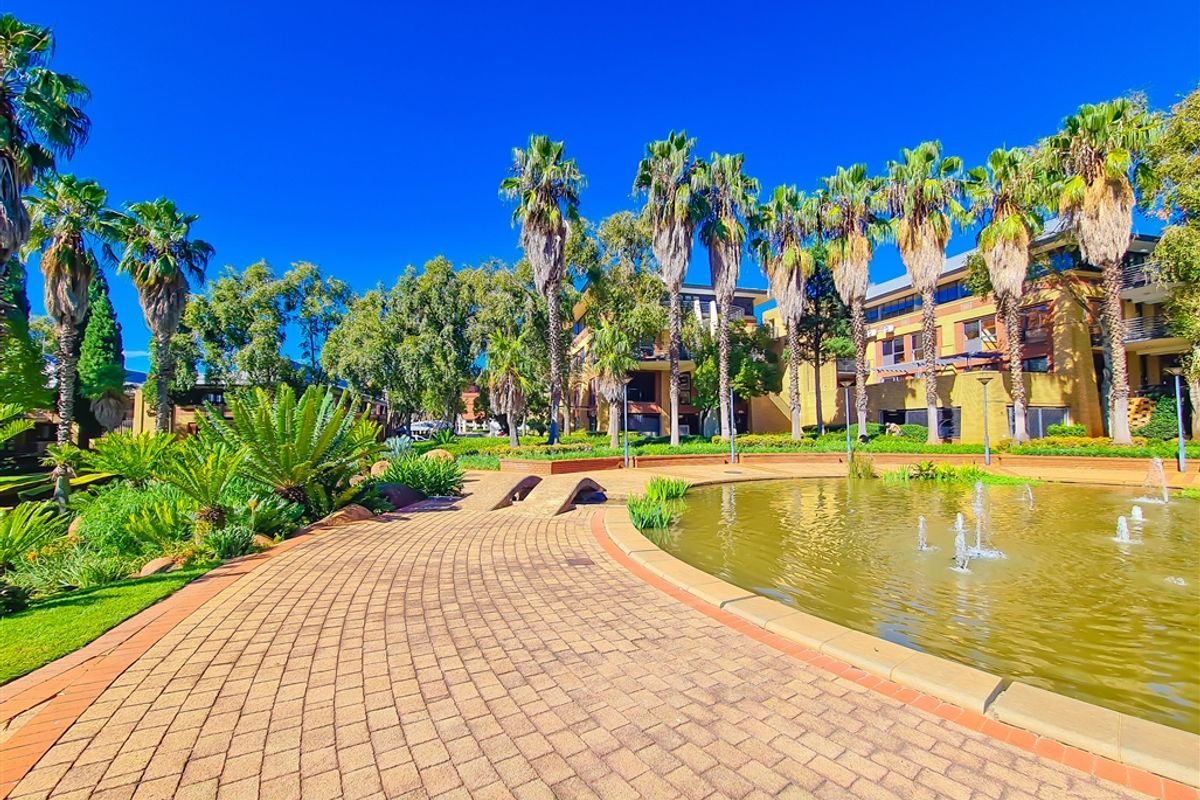 A well-maintained office park exterior featuring a paved walkway, ornamental pond with fountains, and mature palm tree landscaping.