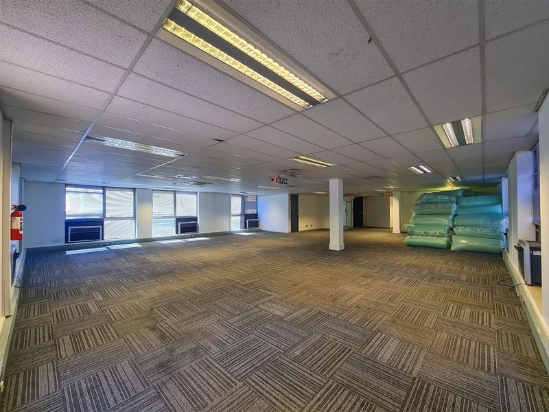 Spacious open-plan office area featuring suspended ceilings, recessed lighting, carpet tiles, and multiple window-mounted air conditioning units.
