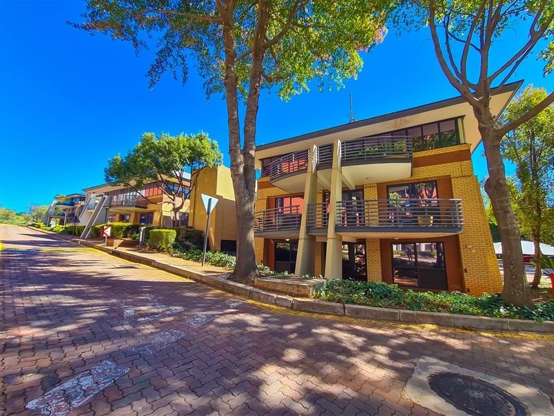 A street-level view of a multi-story office building featuring brick construction, private balconies, and established landscaping along a paved driveway.