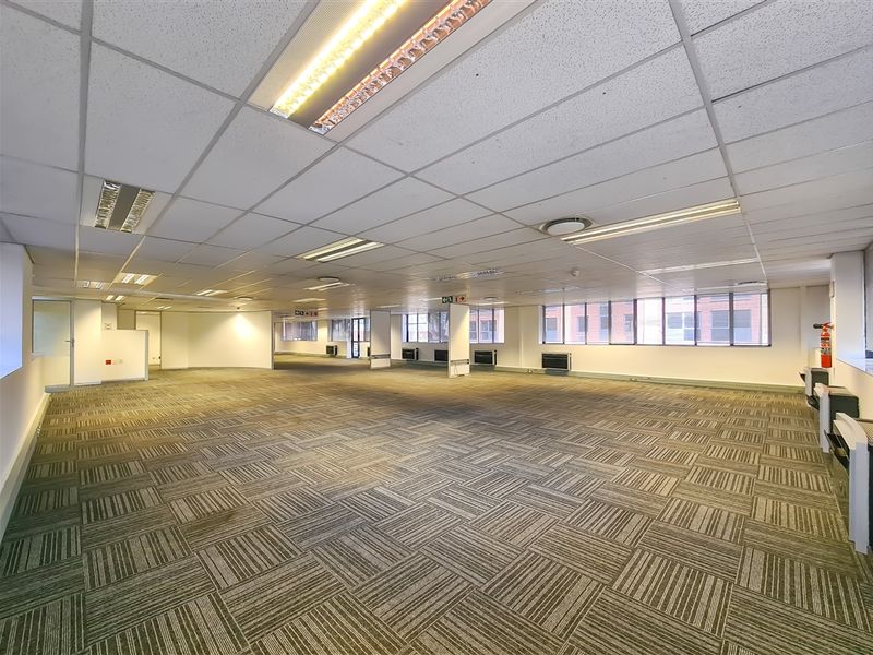 A spacious, open-plan office area featuring carpet tiles, a suspended ceiling with recessed lighting, and large windows providing natural light.