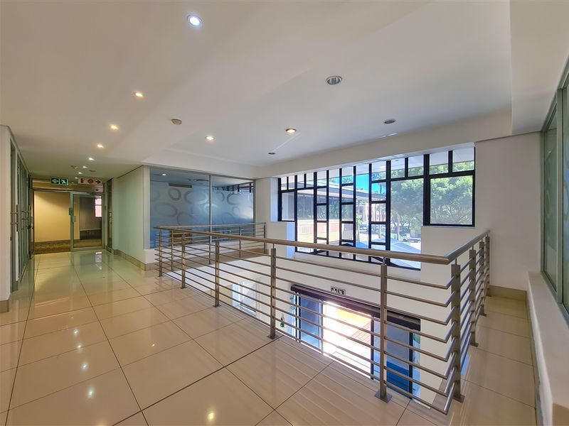 A bright and modern office hallway featuring polished tiled flooring, a sleek metal balustrade overlooking the lower level, and large windows.
