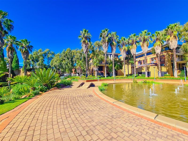 A well-maintained office park exterior featuring a paved walkway, landscaped gardens, and a central water feature with fountains.