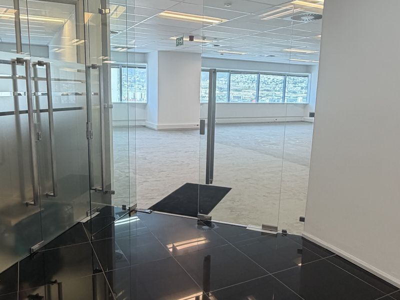 A bright, open-plan office space featuring glass partitions, suspended ceilings with recessed lighting, and a combination of black tile and carpet flooring.