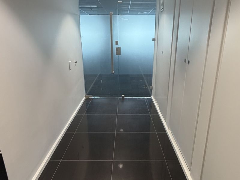 A clean, modern office hallway featuring dark polished floor tiles, recessed lighting, and integrated storage cupboards leading to a glass-partitioned office space.