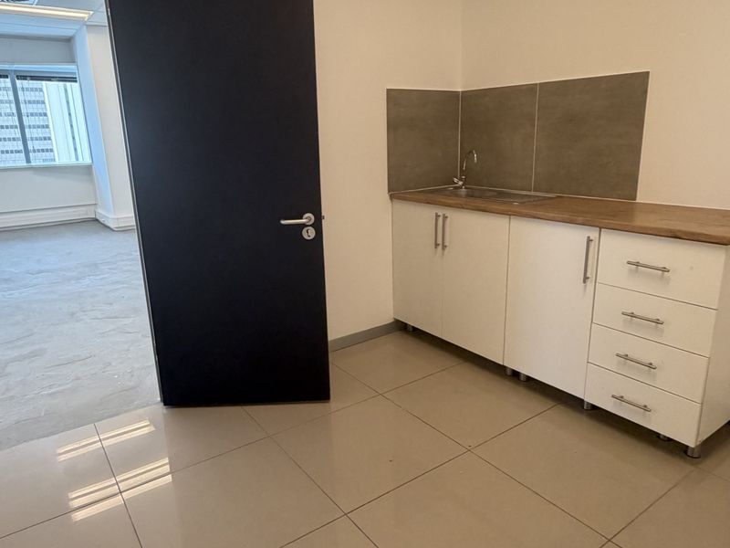 A compact office kitchenette featuring white cabinetry, a stainless steel sink, tiled splashback, and neutral floor tiles in a clean, open-plan office environment.
