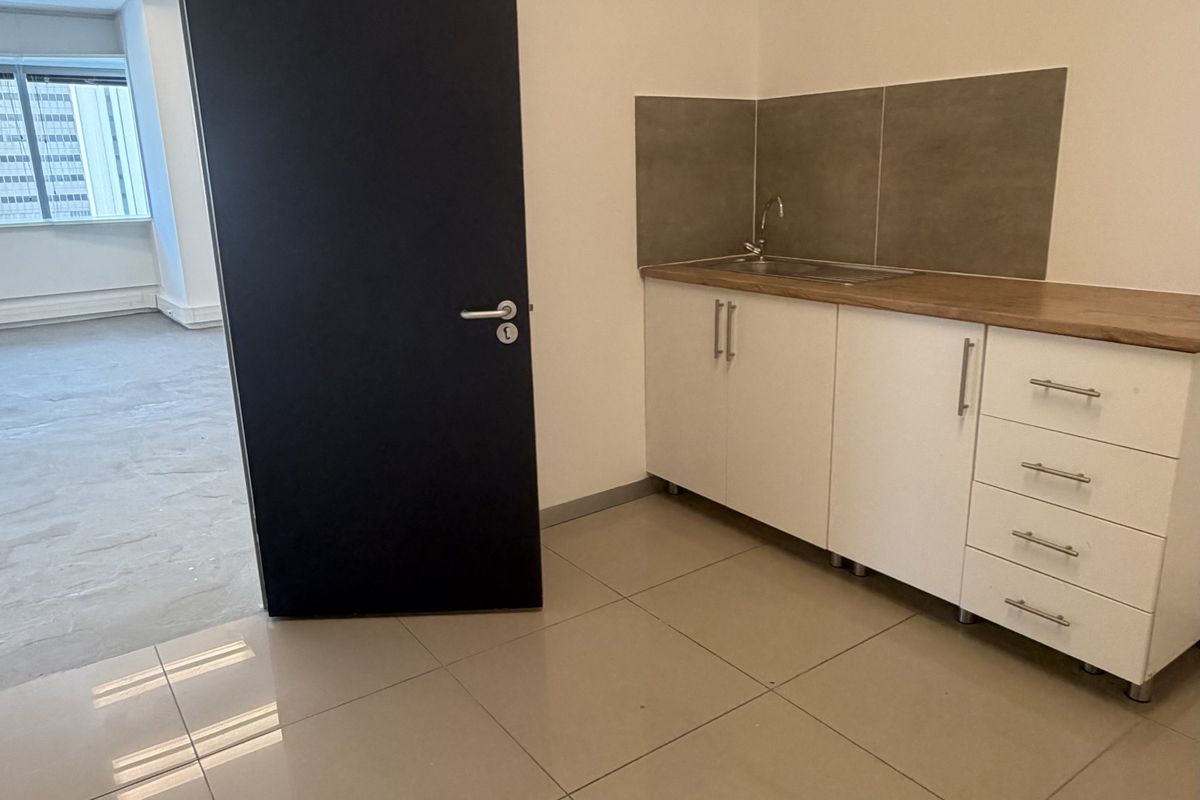 A compact office kitchenette featuring white cabinetry, a stainless steel sink, tiled splashback, and neutral floor tiles in a clean, open-plan office environment.