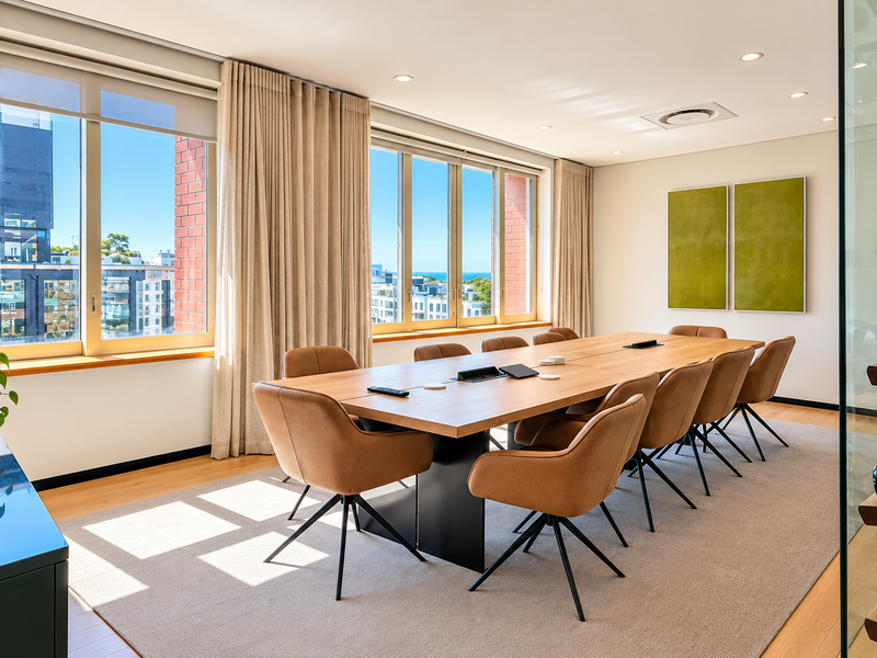 A modern, well-lit office boardroom featuring large windows with city views, hardwood flooring, and integrated ceiling climate control.