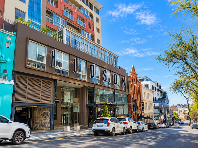 Street-level view of The Hudson building, featuring a modern multi-story facade with large windows, stone detailing, and an integrated parking entrance.