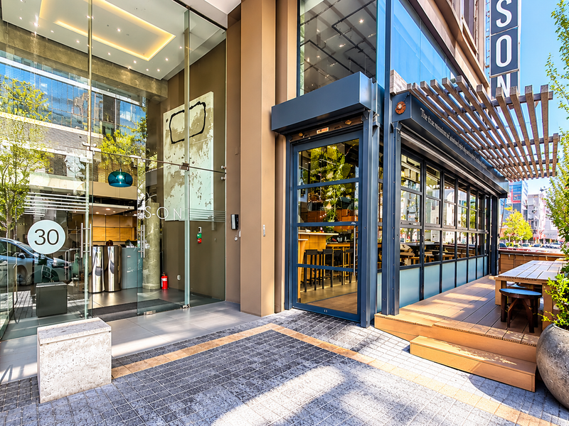 Modern office building entrance featuring a glass facade, timber decking with a pergola, and professional stone paving at the street-level access point.