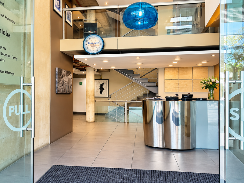 Modern office building entrance featuring a sleek reception desk, glass doors, and a prominent staircase leading to the upper level.
