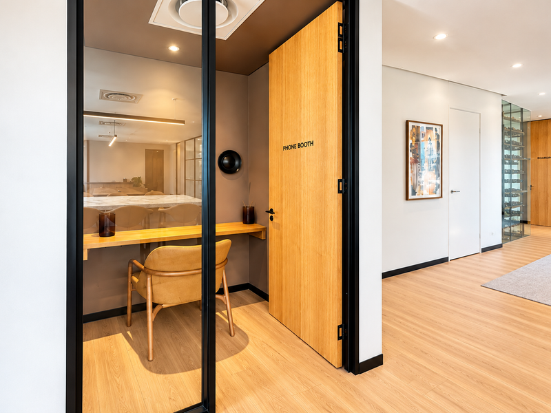 Modern office phone booth featuring a glass partition, built-in desk, and wood laminate flooring, situated within a professional corporate workspace.