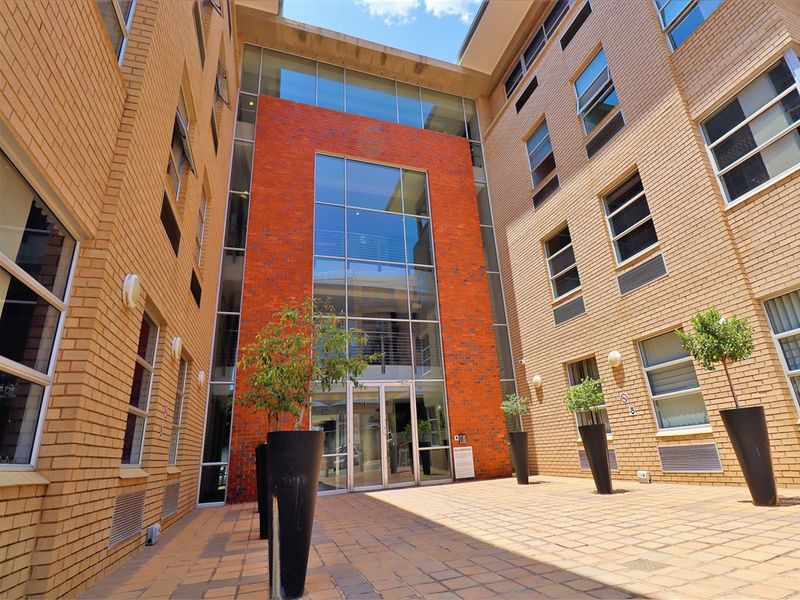 A modern office building courtyard featuring a striking red brick and glass facade with paved flooring and clean architectural lines.