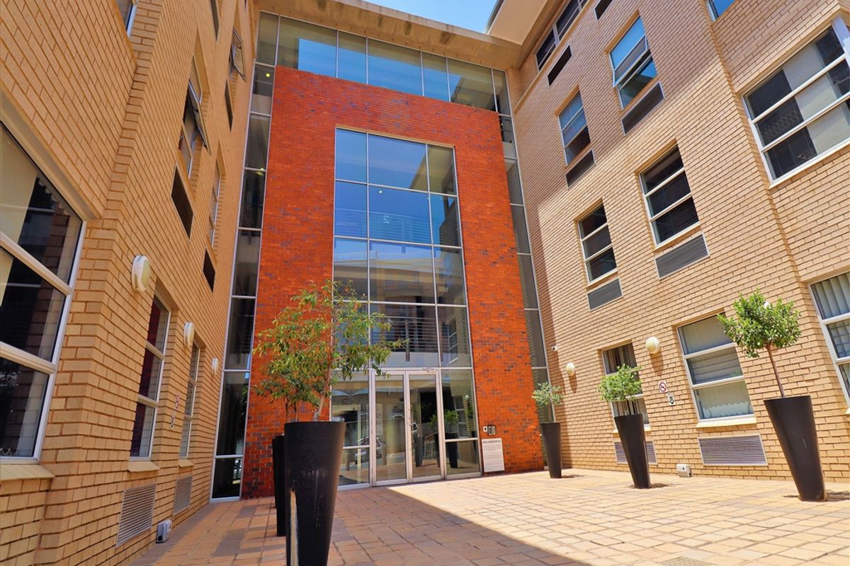 A modern office building courtyard featuring a striking red brick and glass facade with paved flooring and clean architectural lines.