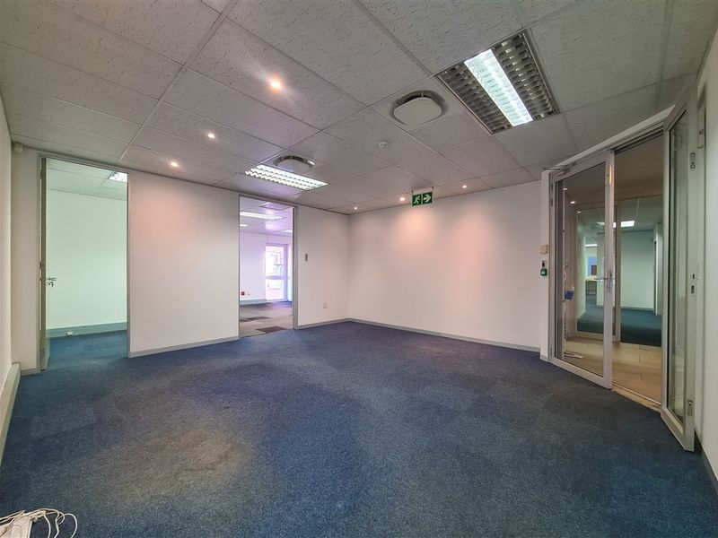 Empty office space featuring a suspended ceiling with recessed lighting, blue carpet flooring, and a glass partition door leading to an adjacent area.