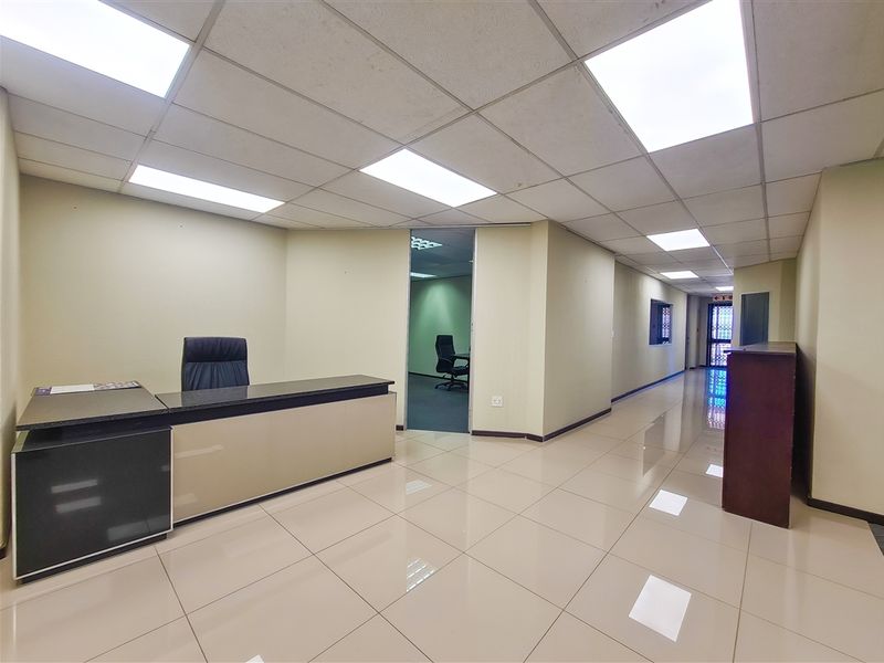 A professional office space featuring tiled flooring, a suspended ceiling with recessed lighting, and an open layout leading to an additional internal office area.