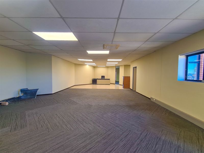 A spacious, empty office area featuring a combination of carpeted and tiled flooring, a suspended ceiling with recessed lighting, and neutral wall finishes.