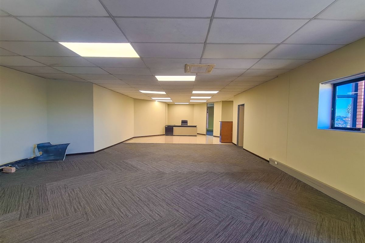 A spacious, empty office area featuring a combination of carpeted and tiled flooring, a suspended ceiling with recessed lighting, and neutral wall finishes.