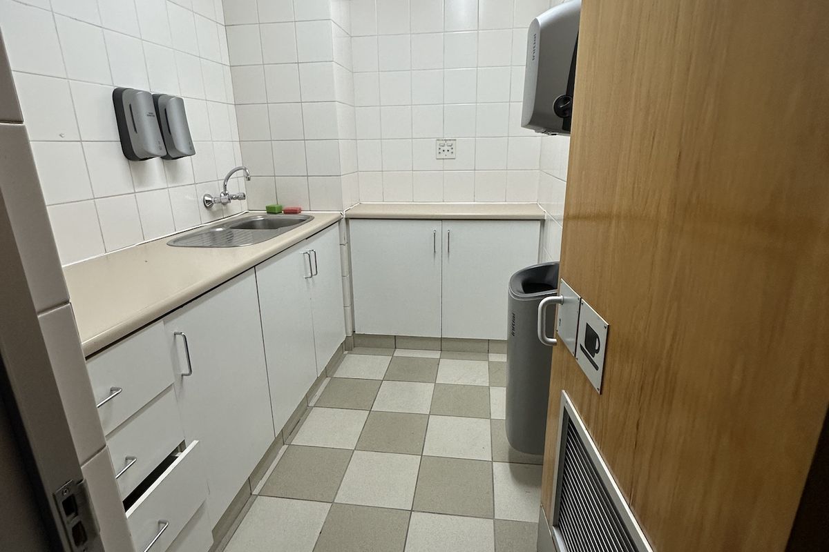 A functional office kitchenette featuring white cabinetry, a stainless steel sink, tiled walls, and a suspended ceiling with integrated lighting.