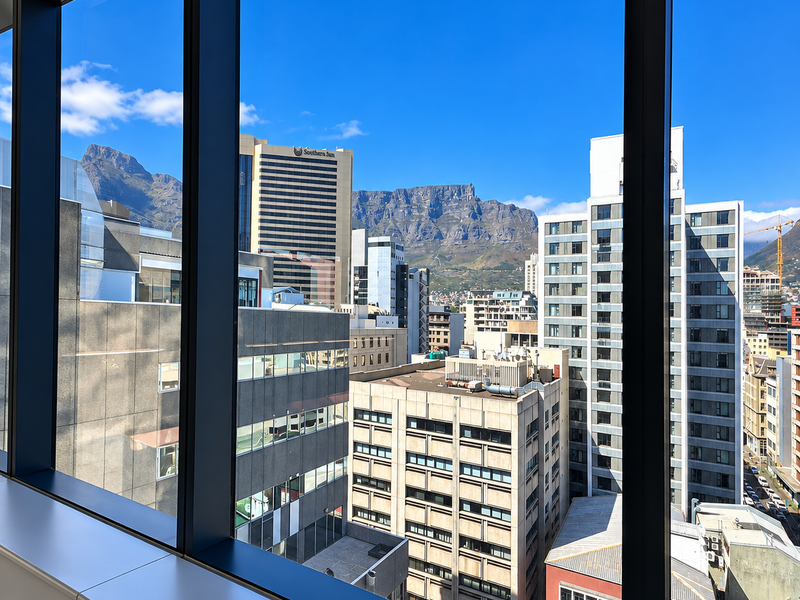 A clear, elevated city view featuring prominent mountain scenery, framed by modern floor-to-ceiling office windows.
