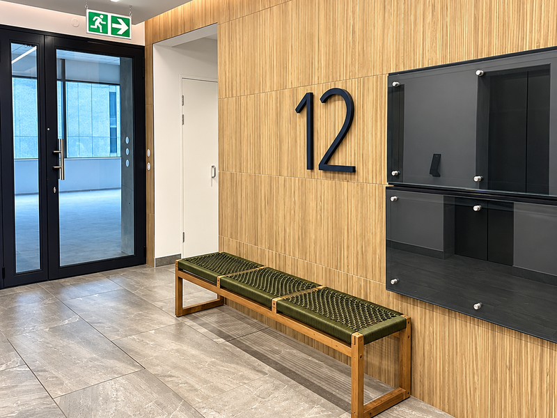 Modern office hallway featuring wood veneer wall paneling, large format porcelain floor tiles, recessed linear lighting, and integrated glass notice boards.
