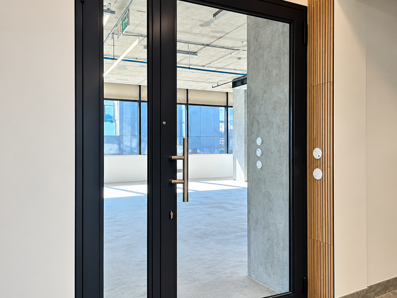 Modern office entrance featuring a sleek double glass door, timber slat wall accents, and polished tile flooring leading into an open-plan workspace.