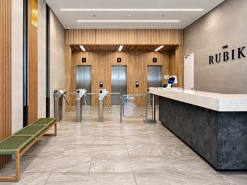 Modern office building lobby featuring a professional reception desk, secure elevator access, and high-end finishes including timber paneling and stone flooring.