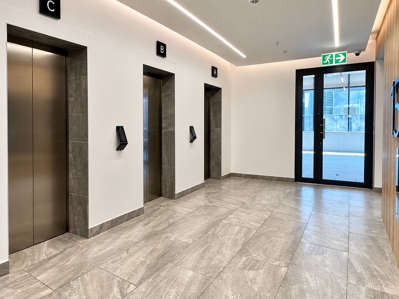 Modern office building elevator lobby featuring high-quality stone tile flooring, three stainless steel elevator banks, and sleek recessed linear lighting.