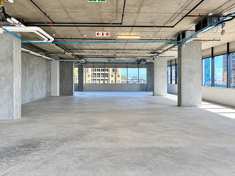 A spacious, open-plan office area featuring an industrial aesthetic with exposed concrete ceilings, fire safety systems, and expansive windows offering natural light.