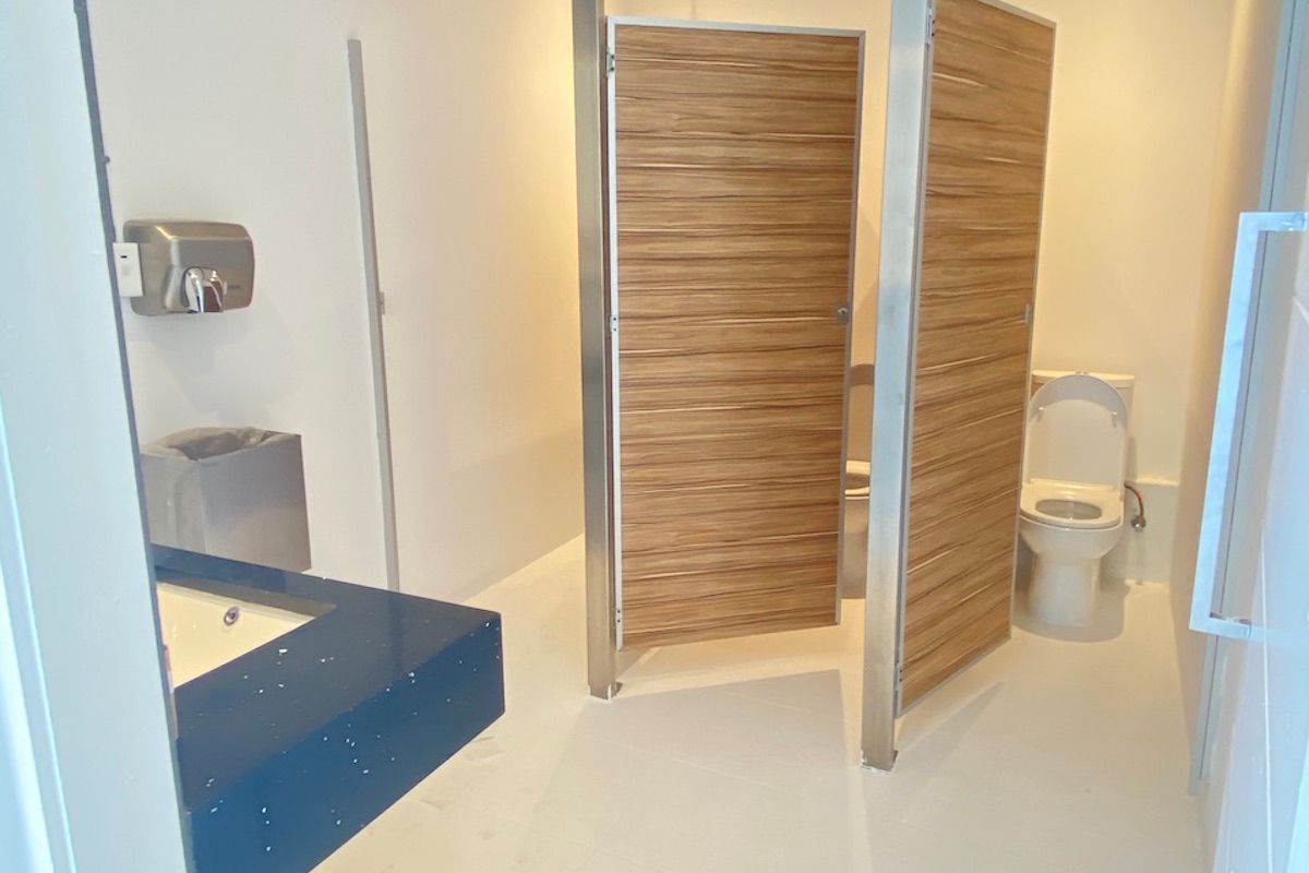 A functional office restroom featuring two toilet cubicles with wood-grain doors, a blue vanity countertop, and a wall-mounted soap dispenser.