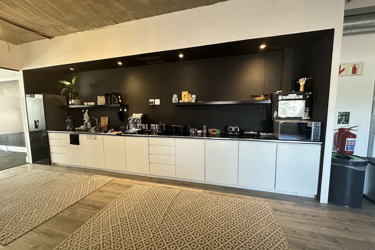 Modern office kitchenette featuring sleek white cabinetry, a dark contrasting backsplash, recessed lighting, and an exposed concrete ceiling.