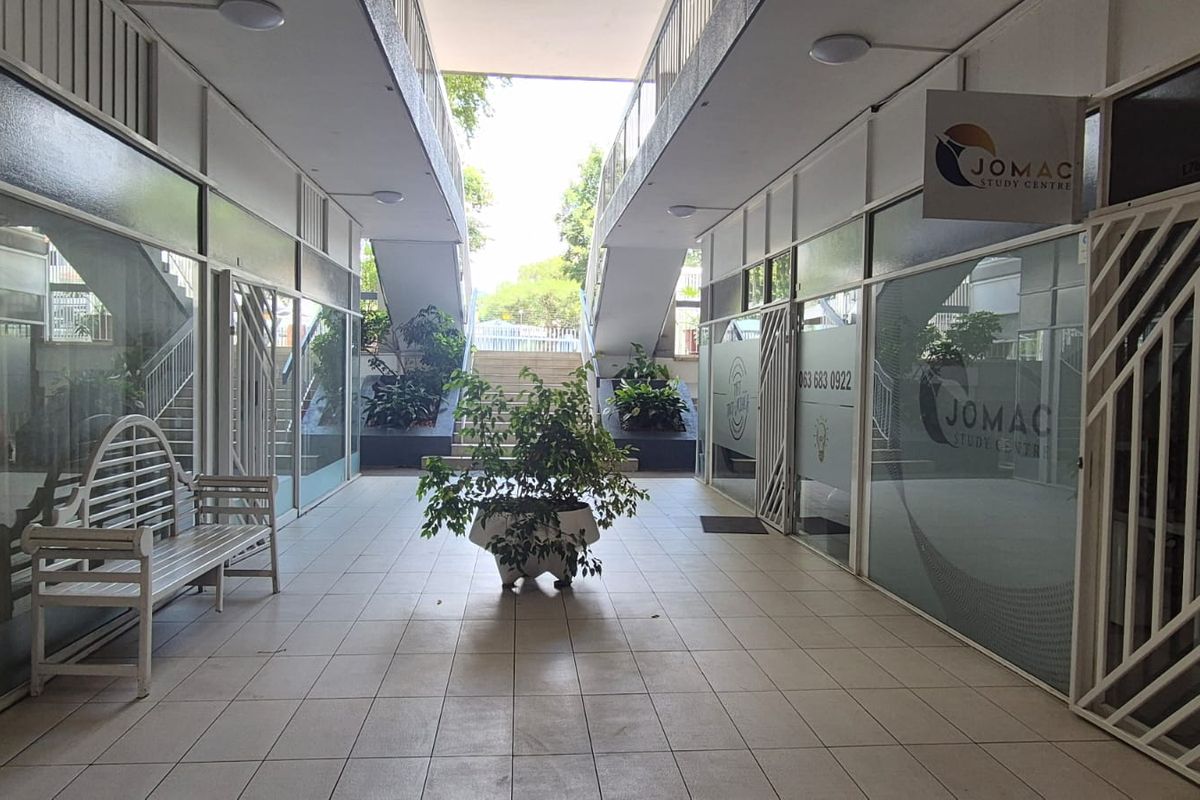 A covered outdoor walkway between office units featuring tiled flooring, glass storefronts, and a central staircase leading to the upper level.