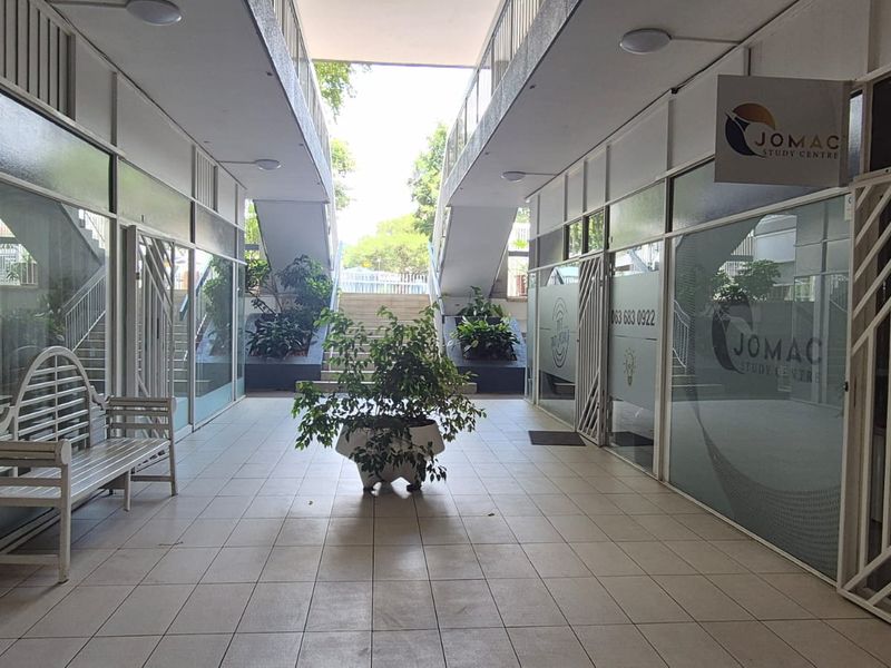 A covered outdoor walkway between office units featuring tiled flooring, glass storefronts, and a central staircase leading to the upper level.