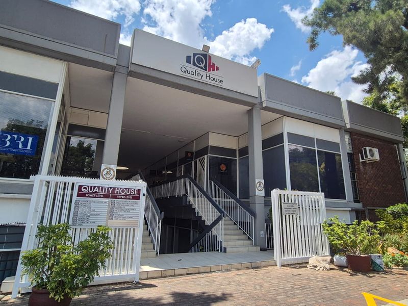 Front elevation of Quality House office building featuring a paved entrance, external signage, metal security gates, and a central staircase leading to the upper level.