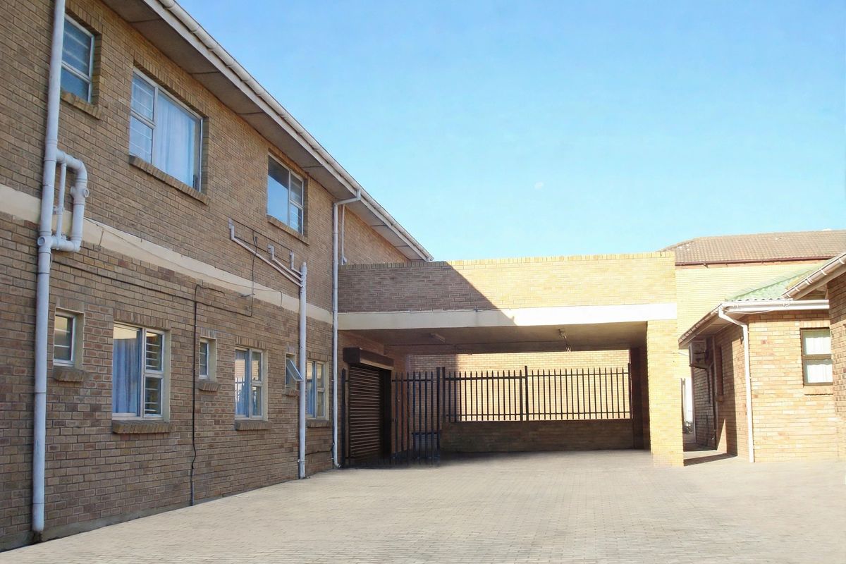 A view of the rear exterior area featuring a face brick building, paved driveway, and a covered carport with security gate access.