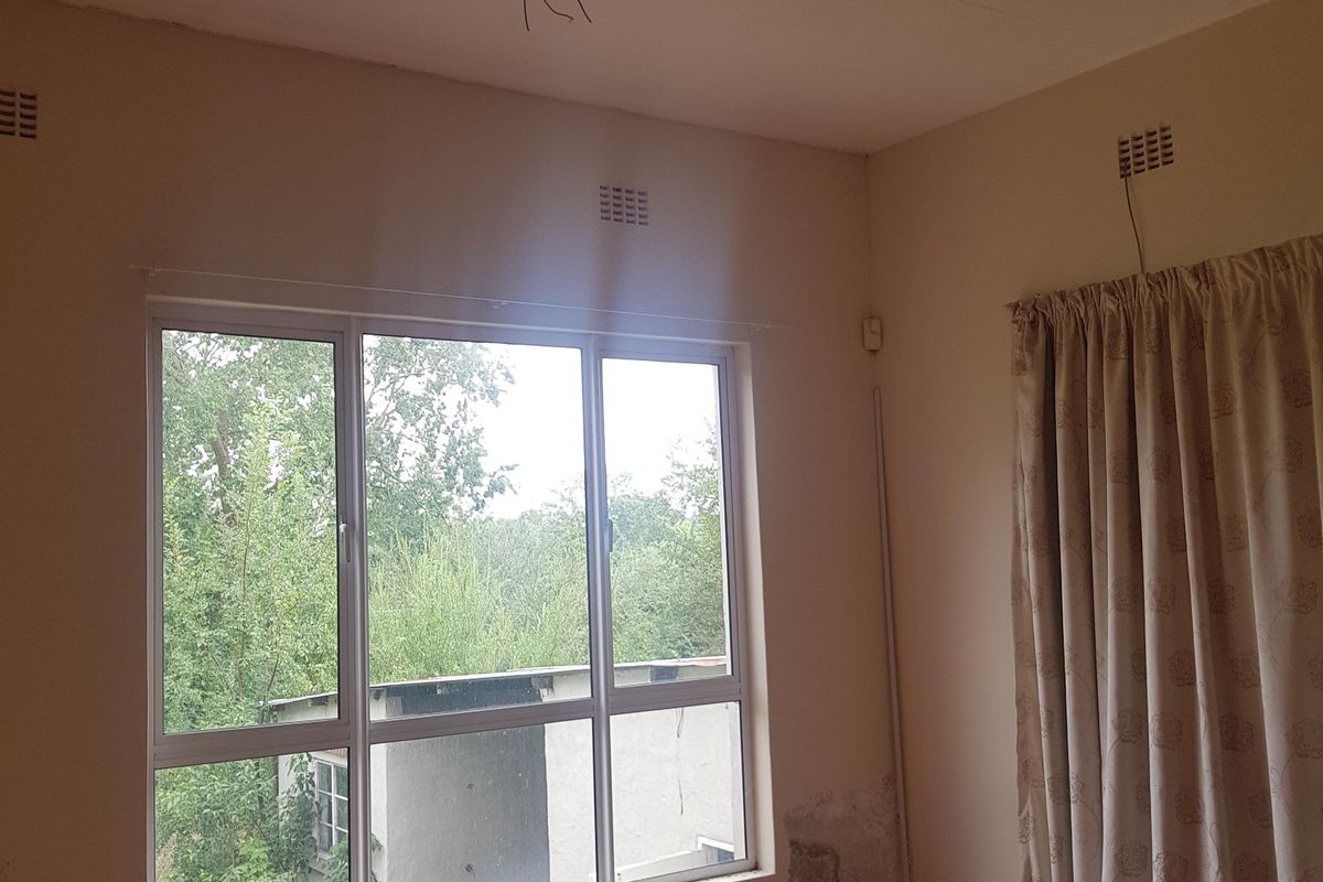 An empty bedroom featuring tiled flooring, a ceiling fan, and aluminium windows, currently requiring wall repairs and maintenance due to visible damp and peeling paint.