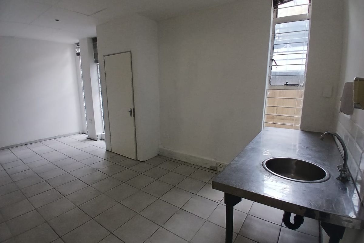 A basic office kitchenette area featuring a stainless steel sink unit, tiled flooring, and large windows with security bars.