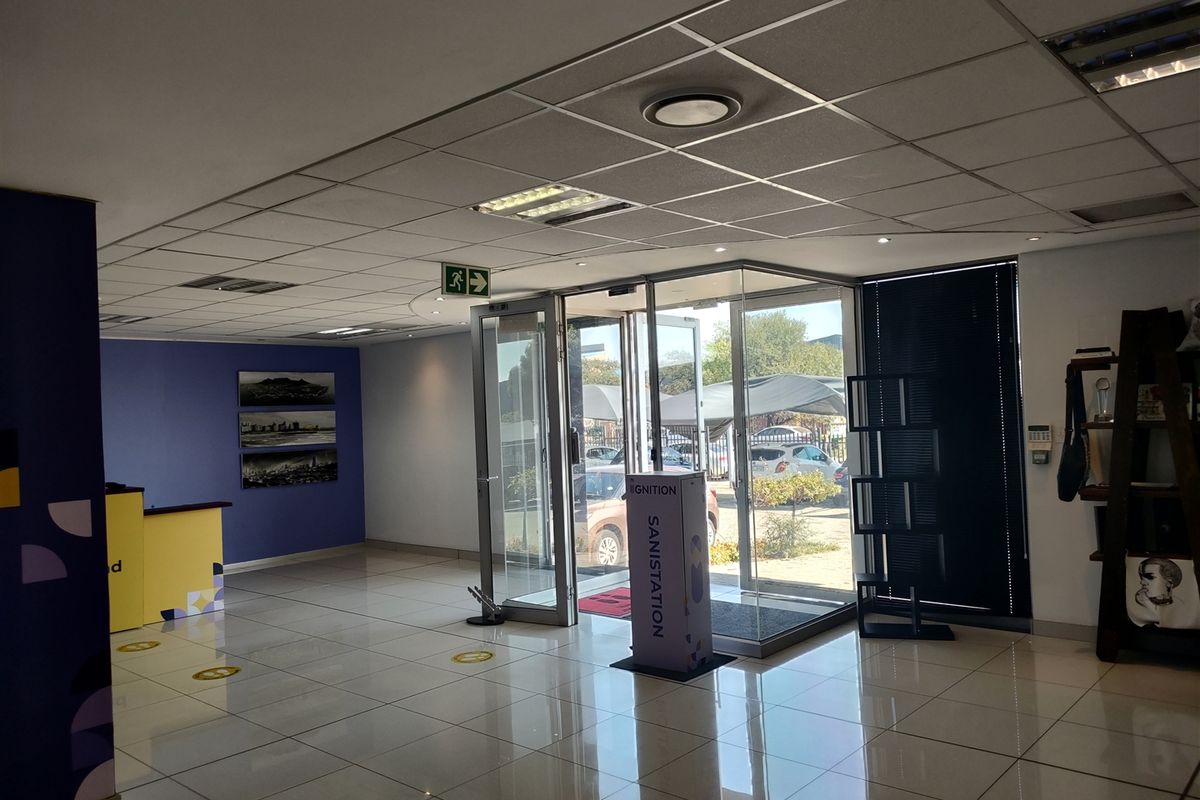 A bright and spacious industrial office entrance featuring tiled flooring, a suspended ceiling with recessed lighting, and glass double doors leading to the exterior.