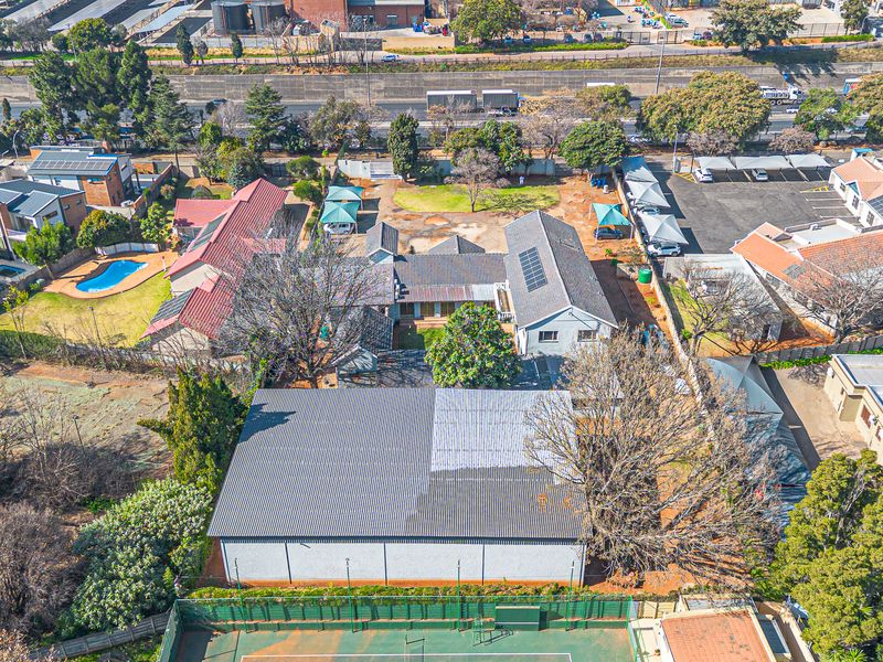 Aerial view of a commercial property featuring a large warehouse structure, tennis court, paved parking with shade ports, and surrounding landscaped grounds.