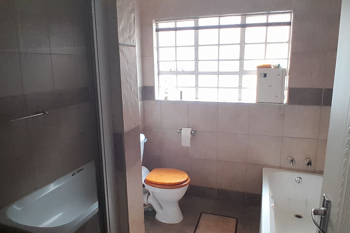 A functional bathroom featuring tiled walls and flooring, a built-in bathtub, and a toilet, with natural light provided by a window with security bars.
