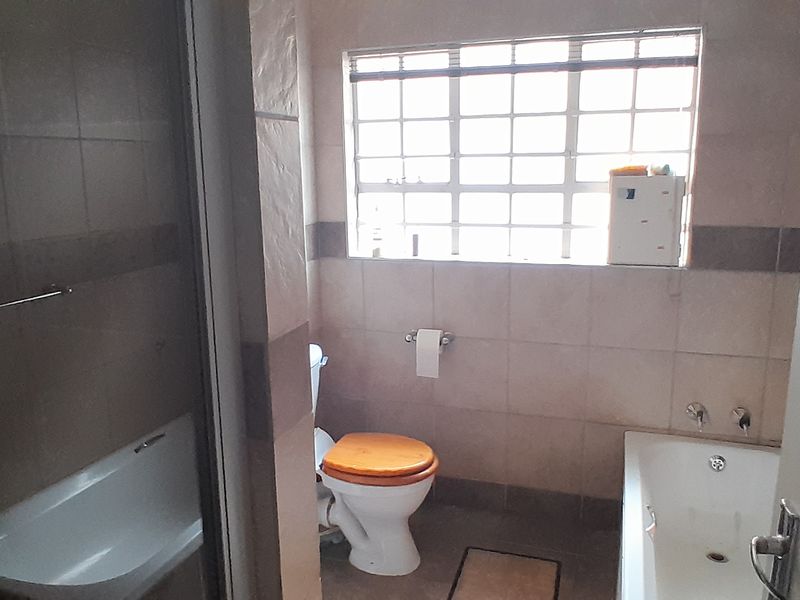A functional bathroom featuring tiled walls and flooring, a built-in bathtub, and a toilet, with natural light provided by a window with security bars.