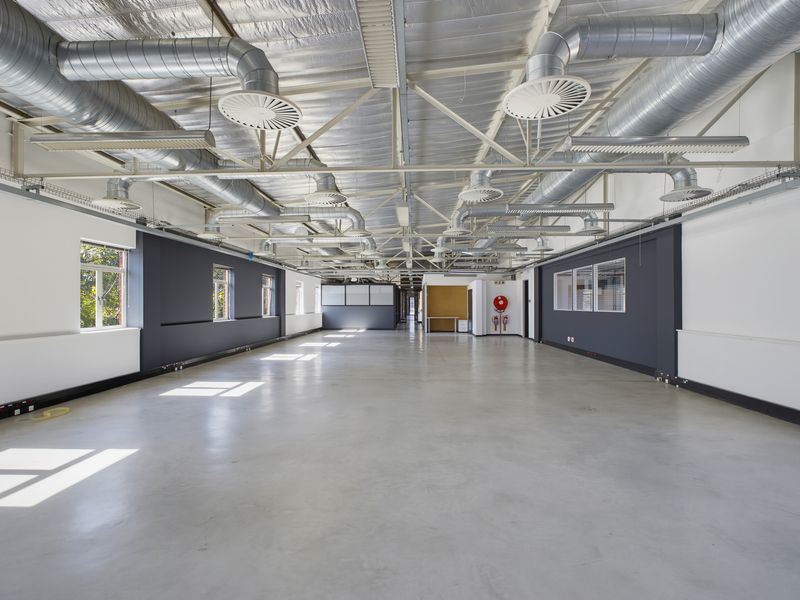 Spacious open-plan office area featuring high ceilings with exposed industrial ductwork, polished concrete floors, and ample natural light from side windows.